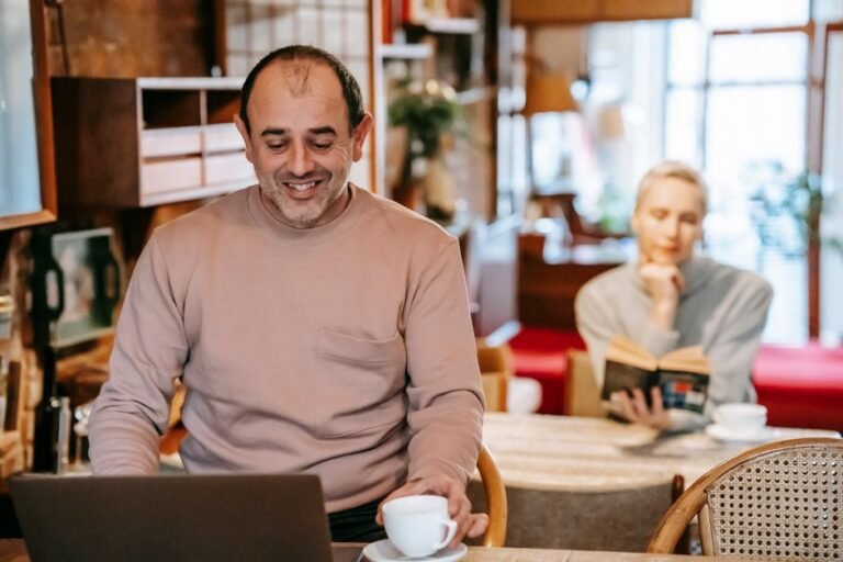 Positive middle aged ethnic male freelancer drinking coffee and working remotely in laptop near focused wife reading book at home