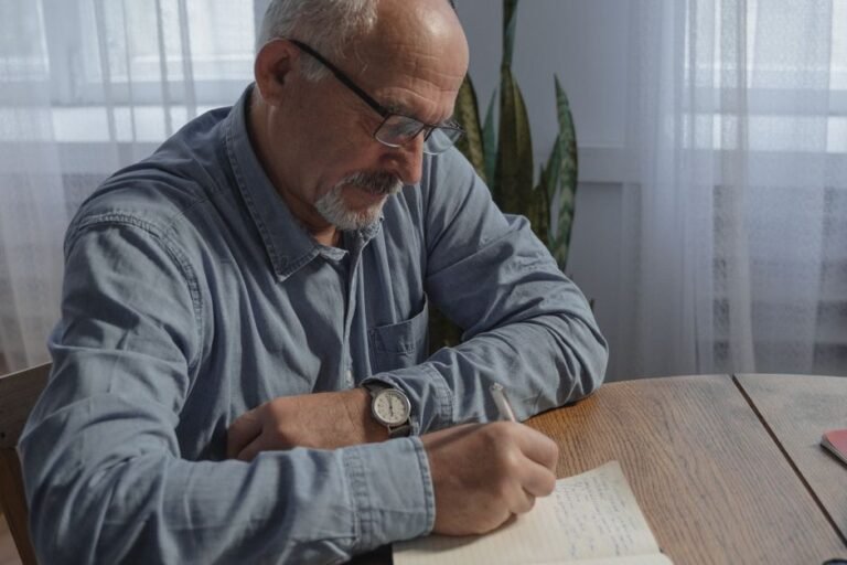 Elderly man writing notes at a table, engaged in remote work at home.